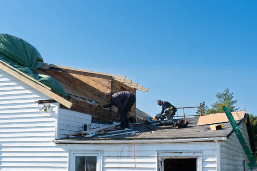 Roof repair workers installing shingles under a clear blue sky. Safety equipment visible.