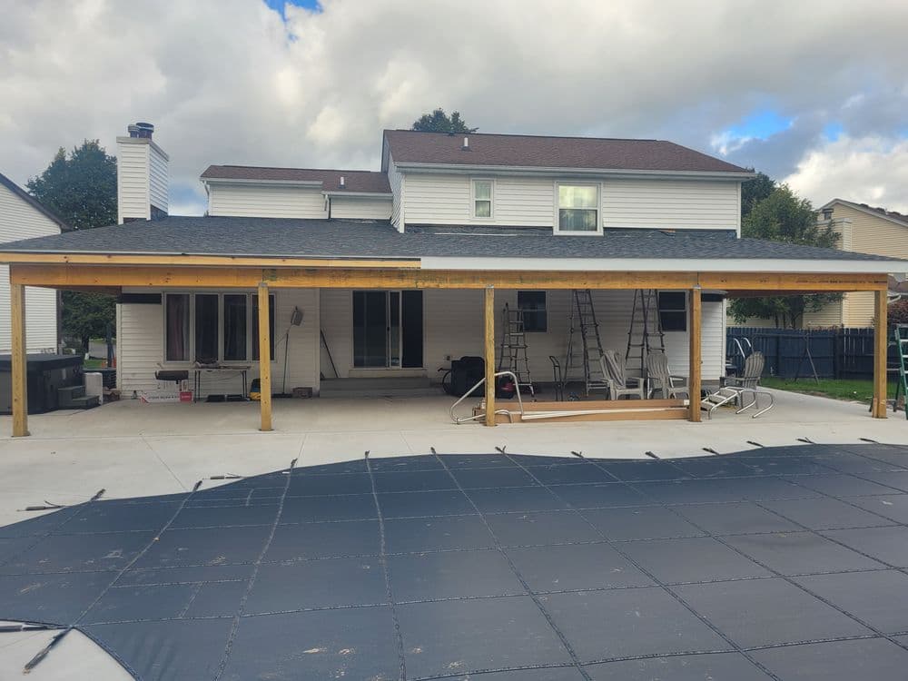 Backyard patio with wooden pergola, concrete floor, and house in background.