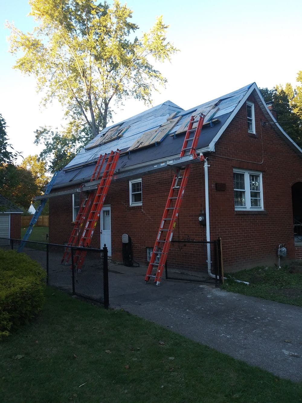 Red brick house with ladders leaning against roof, undergoing repair during sunny day.