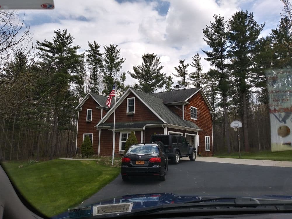 Charming red-shingled house with American flag, parked cars, and tall pine trees in driveway.