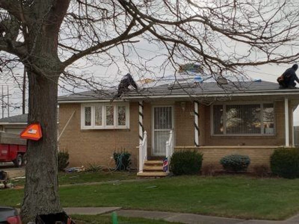 Roofing contractors working on a residential home with a tree in the foreground.