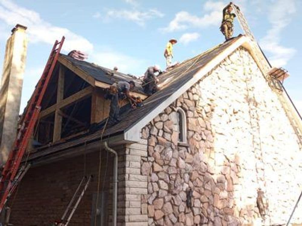 Roofers repairing a stone house, with ladders and tools visible against a blue sky.