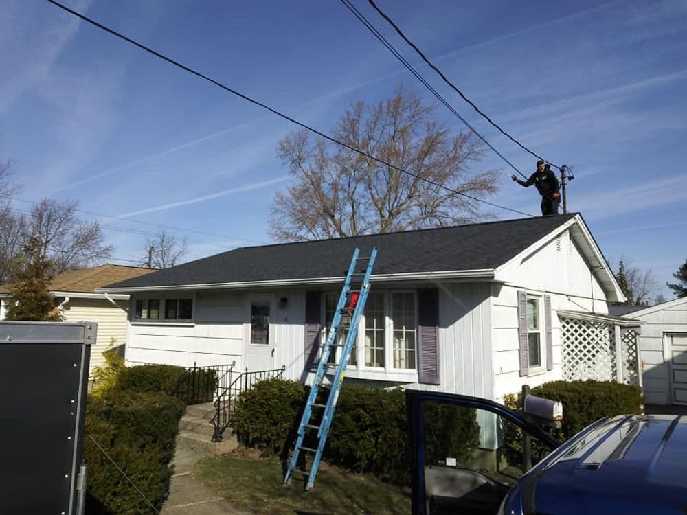 Roofer applying shingles on a house with a ladder and blue sky in the background.
