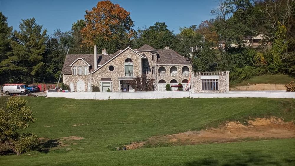Stone mansion with arched windows set on a green landscape, surrounded by trees in autumn.