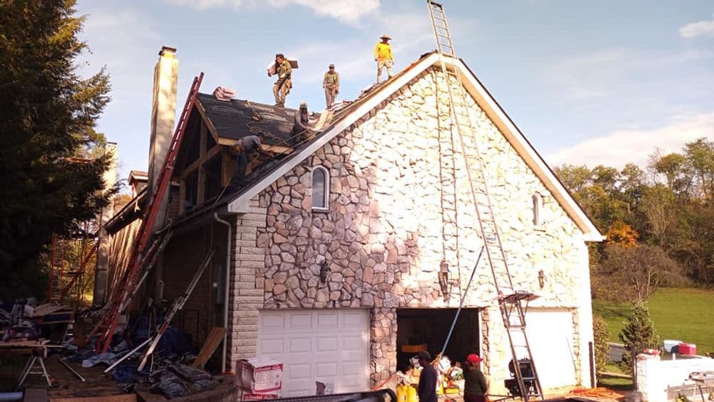Workers repairing a stone house roof with ladders and equipment in a scenic outdoor setting.