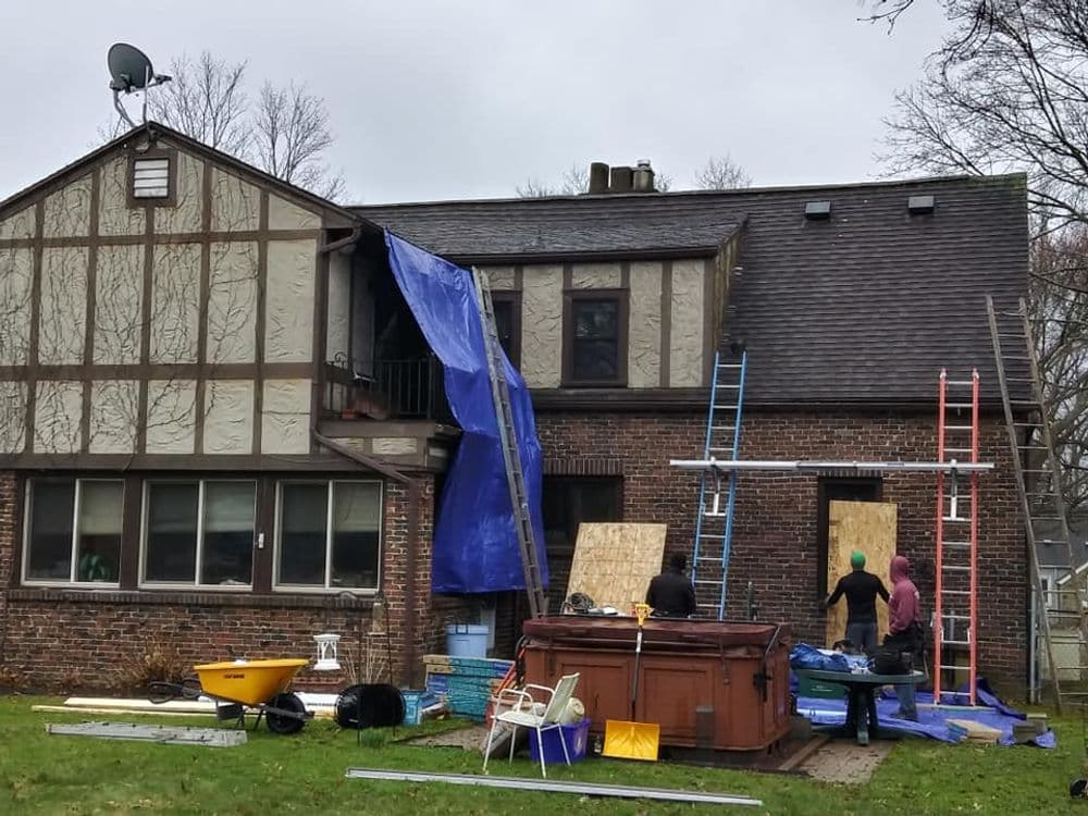 Home exterior renovation with blue tarp, workers, ladders, and a hot tub in the yard.