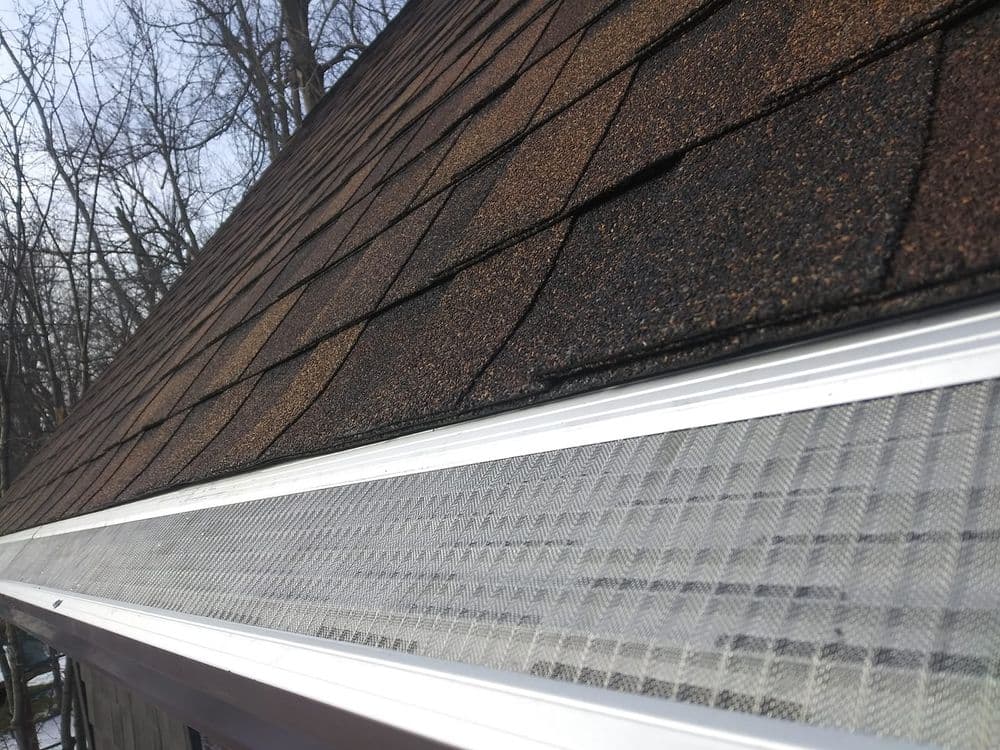 Close-up of a shingled roof with a metal gutter guard and trees in the background.