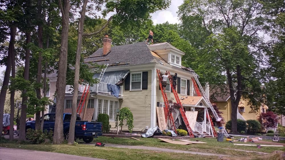 Workers repairing a house roof with ladders, surrounded by trees on a sunny day.