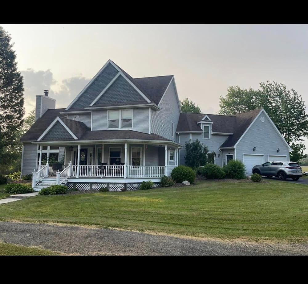 Large charming gray house with a porch, well-maintained lawn, and trees in the background.