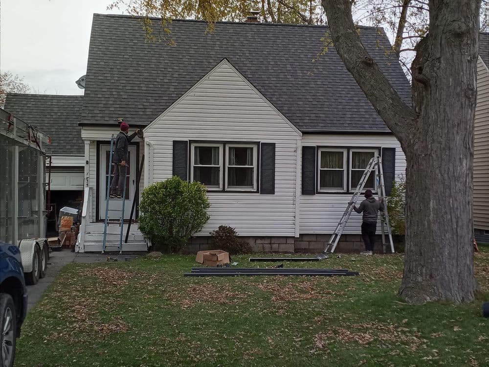 Workers on ladders performing exterior maintenance on a house with a gray roof and white siding.