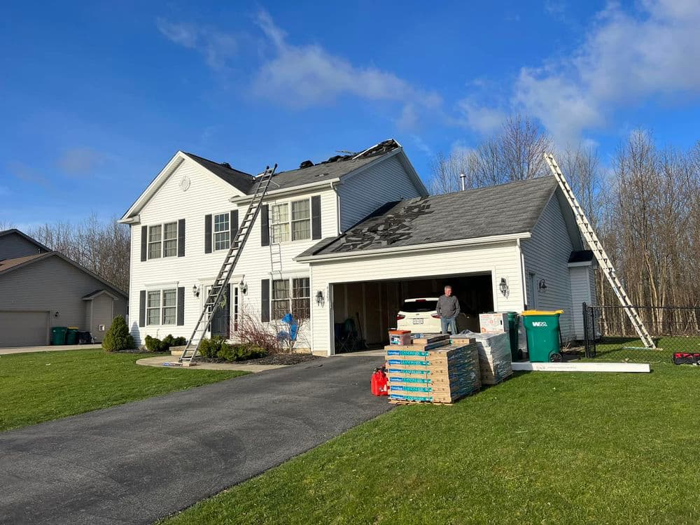 Roofing installation on a suburban home with ladders, materials, and a worker on site.
