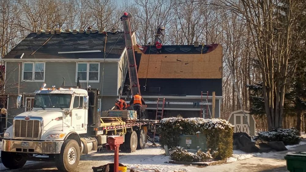 Roof replacement in progress with workers and crane, snowy residential area.