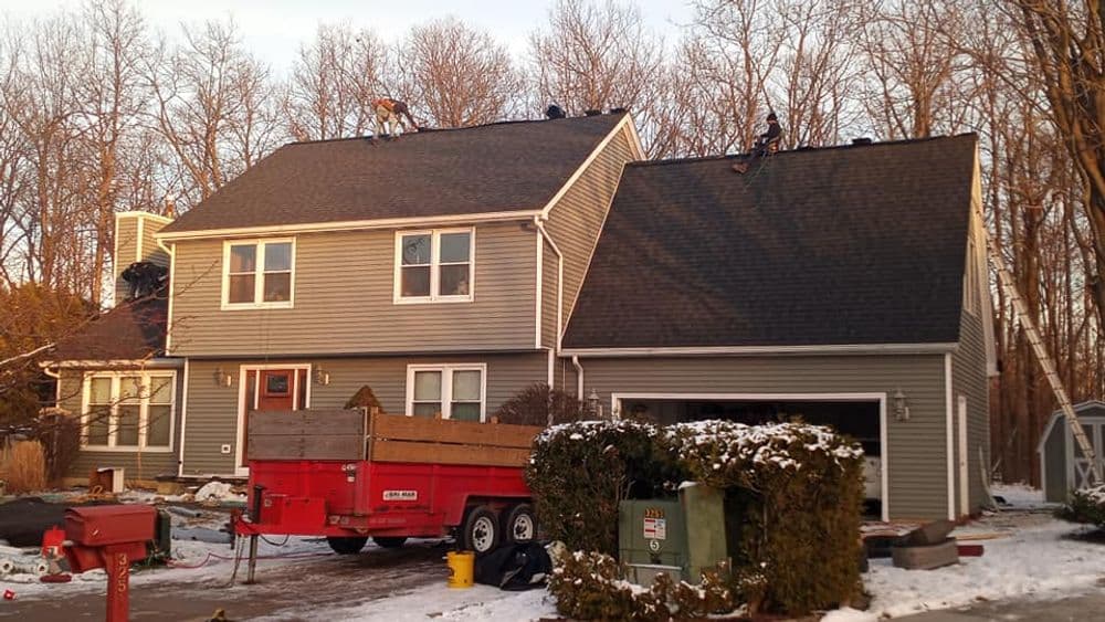 Roof maintenance on a gray house in winter, with workers on the roof and snow in the yard.