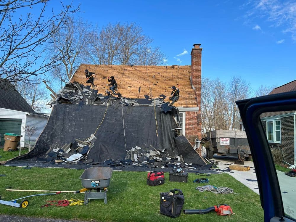 Workers removing shingles from a house roof, debris covered with a tarp in front yard.
