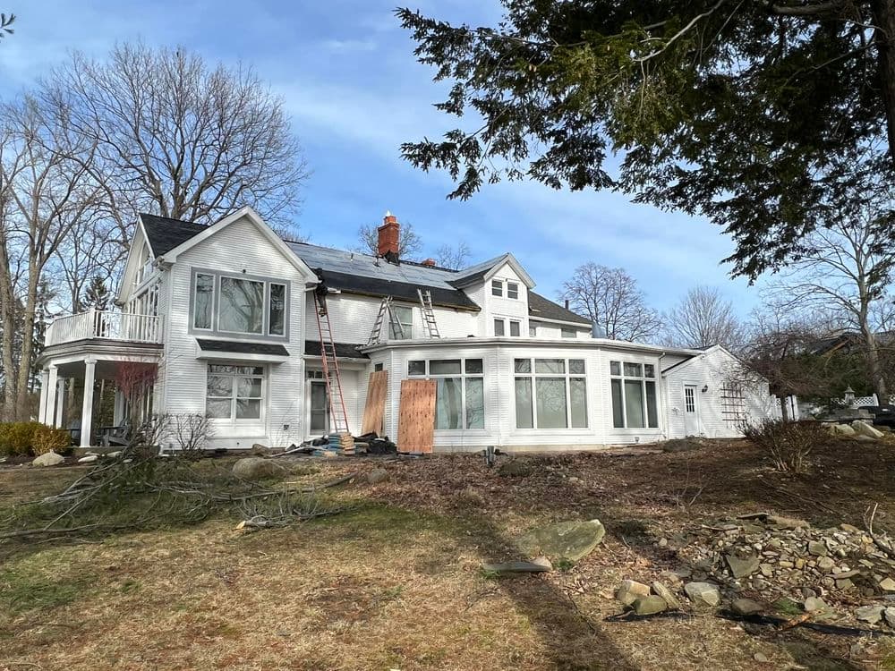 White house undergoing renovation with ladders and solar panels in a wooded area.
