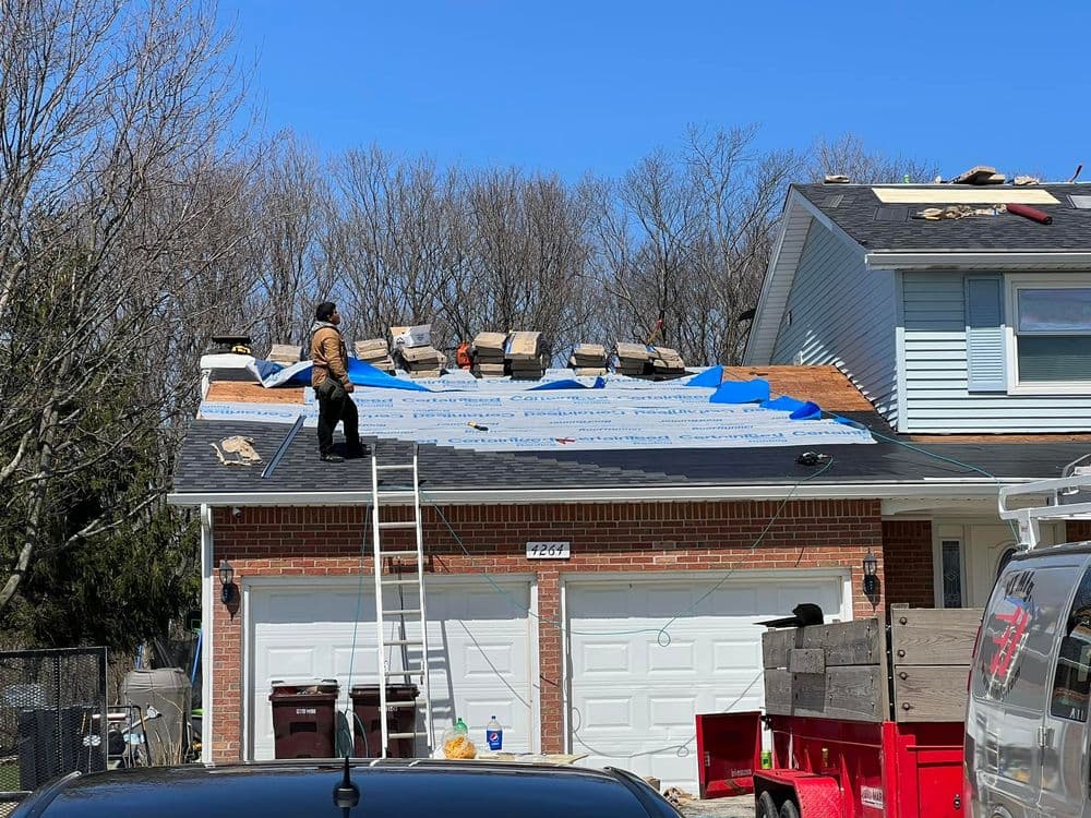 Roofer installing shingles on residential roof with blue underlayment on a sunny day.