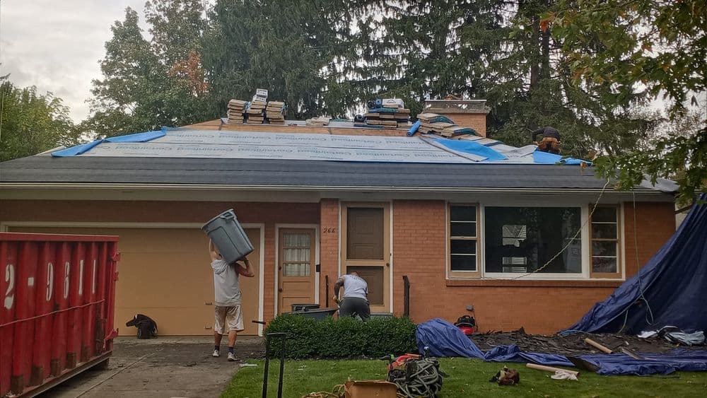 Roofing installation in progress on a brick house with workers and debris visible.