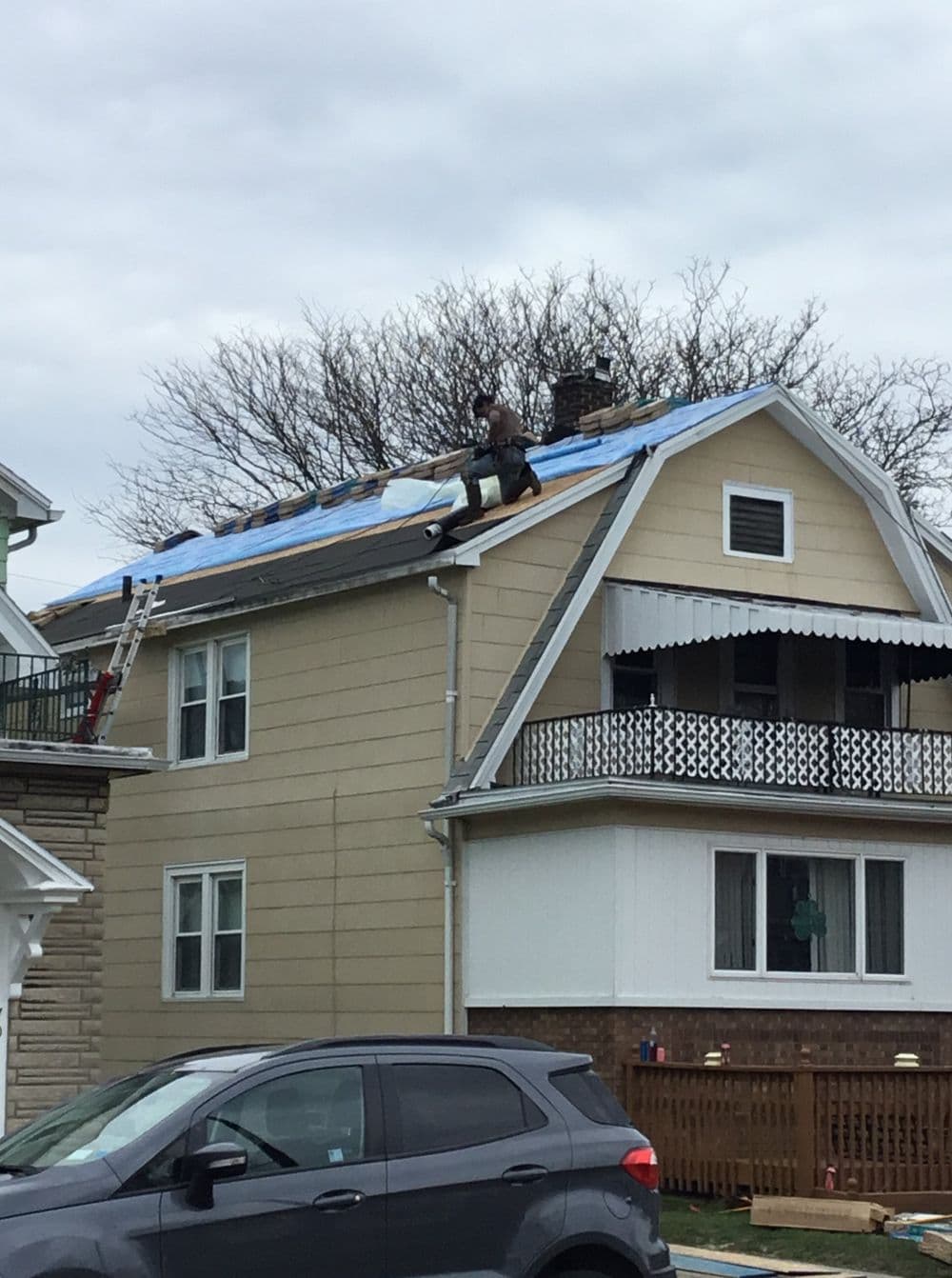 Man repairing a roof on a beige two-story house with trees and a cloudy sky.