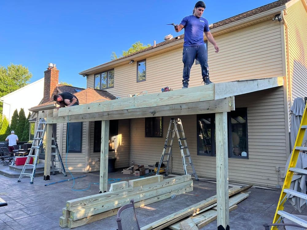 Workers constructing a wooden deck on a home, with ladders and materials visible on-site.