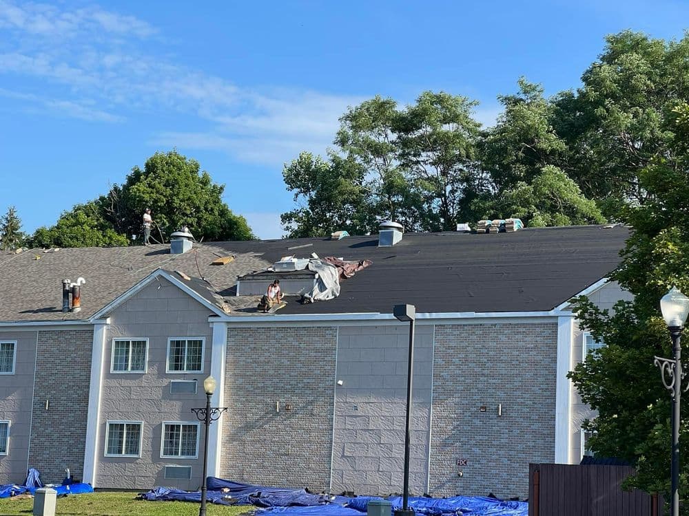 Roofing workers installing new shingles on a residential building with blue tarps below.