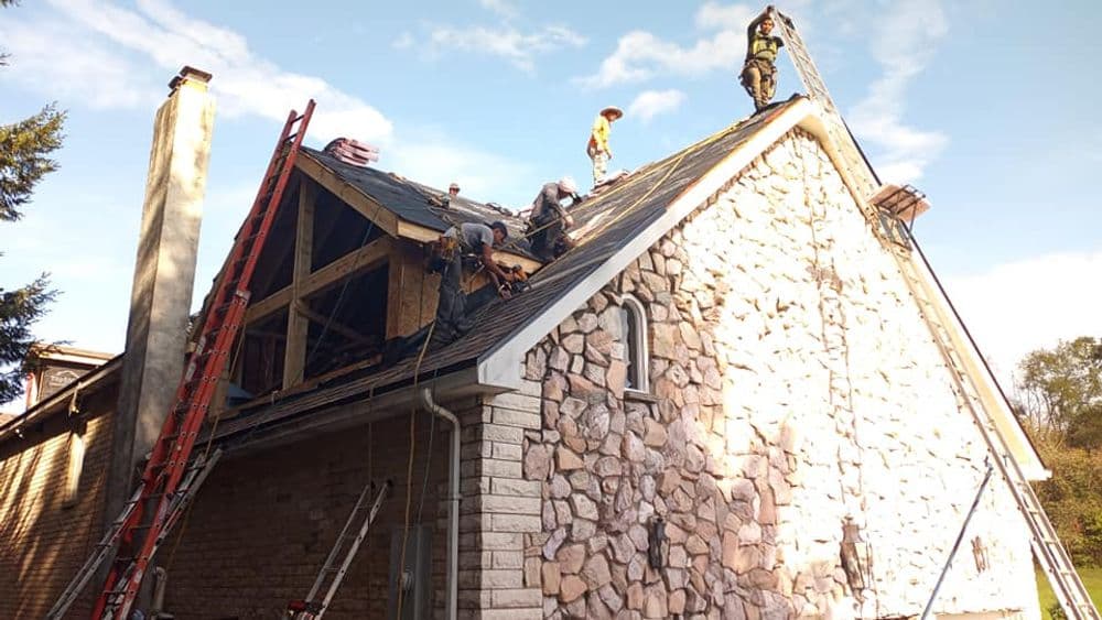 Roofing contractors repairing a stone house under blue sky with ladders and scaffolding.