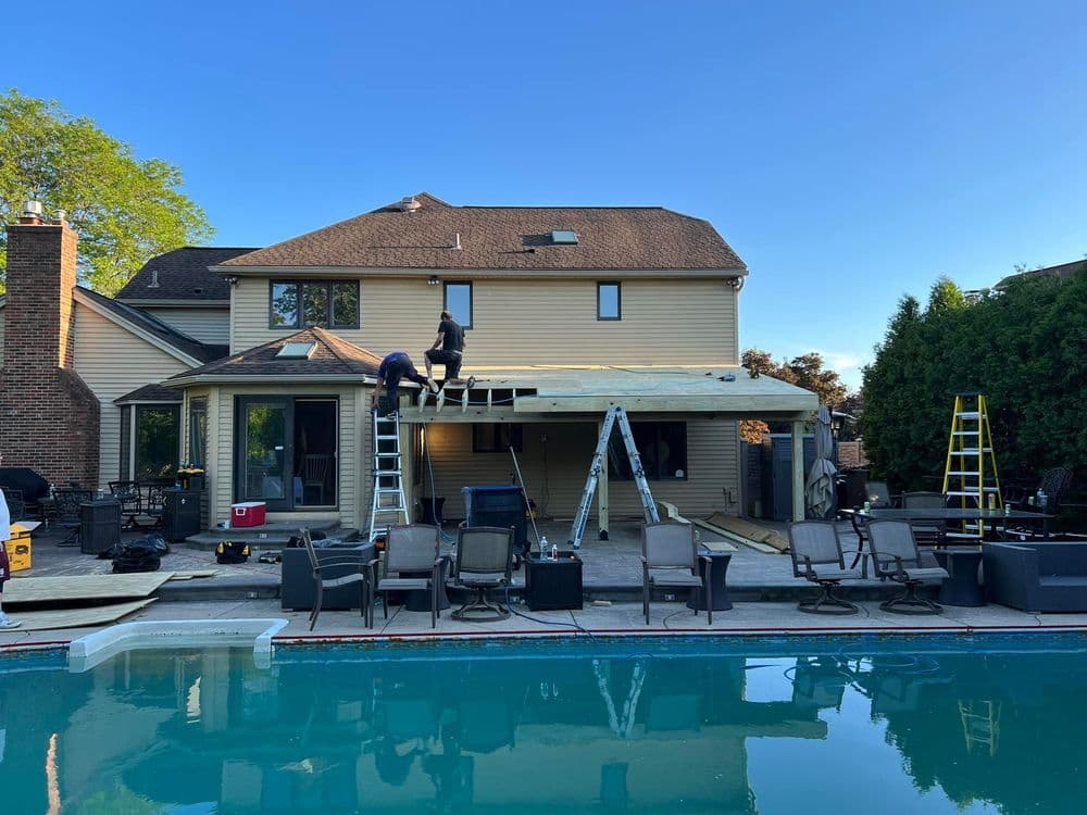 Workers constructing a patio cover behind a house by a swimming pool on a sunny day.