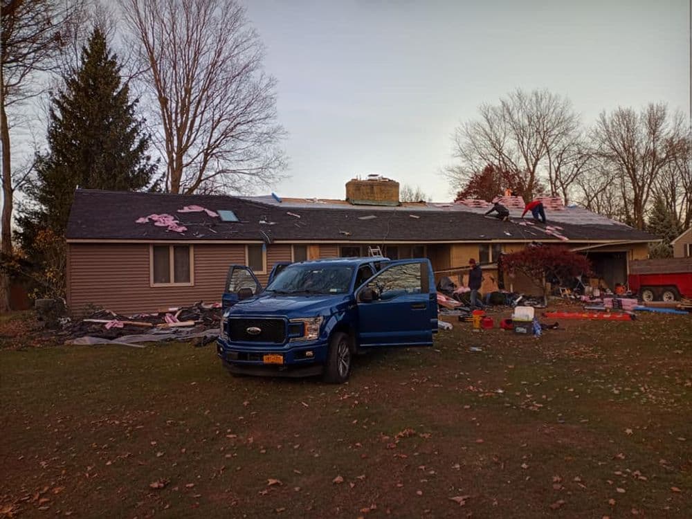 Blue pickup truck parked outside a house with workers installing a new roof on a sunny day.